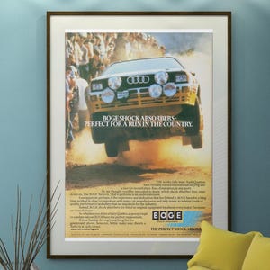 May include: A framed advertisement featuring a yellow Audi Quattro rally car mid-air, with the text "BOGE SHOCK ABSORBERS - PERFECT FOR A RUN IN THE COUNTRY." The poster is displayed in a room with a yellow chair and a glass coffee table.