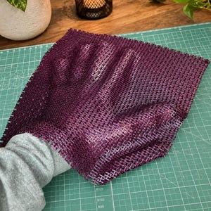 May include: A square, deep purple, flexible mesh sheet with a grid-like texture. The material is semi-transparent and features a repeating pattern of small, interconnected squares. It is shown being held, revealing a hand shape underneath.