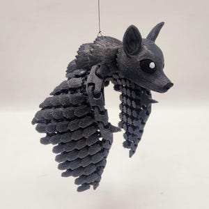 May include: A black, 3D-printed fox ornament with articulated wings and a detailed, textured surface. The fox has pointed ears and large eyes. The ornament is suspended by a thin black string, suitable for hanging as a decoration.