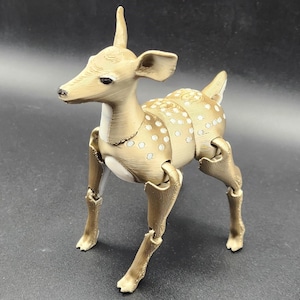 May include: A tan articulated deer figurine with white spots on its back and sides. The deer has a small, pointed antler and is posed with its legs extended. The figurine is made of a light-colored material and is set against a dark background.