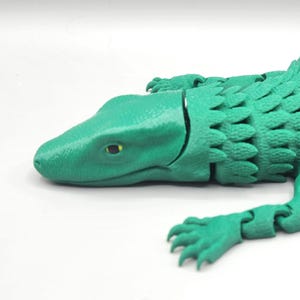 May include: A green articulated lizard toy. The toy is a vibrant shade of green, with a segmented body, legs, and tail. The lizard is lying down, showcasing its detailed, textured design. The eyes are a contrasting yellow-green.
