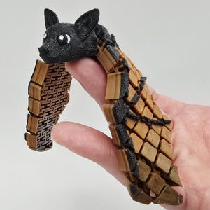 May include: A 3D-printed articulated cat toy in black and brown. The cat's head is black with glittery eyes, and the body is a segmented design in shades of brown and black. The toy is held in a hand.
