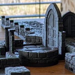Puede incluir: Un conjunto modular de terreno de mazmorra gris para juegos de mesa. El conjunto incluye varios componentes como muros, arcos, pilares y un pozo, todos diseñados para crear un entorno detallado e inmersivo para juegos de rol. Las piezas están dispuestas sobre una superficie de madera.