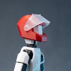 Può includere: Action figure bianca articolata con casco rosso e visiera trasparente. La figura ha guanti neri e dettagli sulle articolazioni. La figura è in posa con un braccio teso.