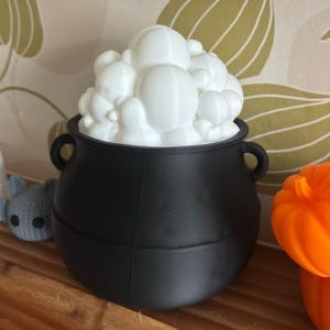 May include: A black cauldron filled with white, puffy shapes, possibly representing bubbles or foam. The cauldron has two handles. Beside it are a small crocheted ghost and bat, and an orange pumpkin. The items are on a wooden shelf.