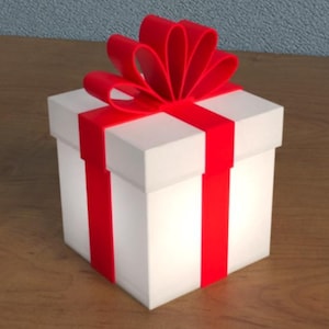 Puede incluir: Una caja de regalo blanca con una cinta y un lazo rojo brillante. La caja tiene rayas verticales rojas y está sobre una superficie de madera. La caja de regalo está iluminada.