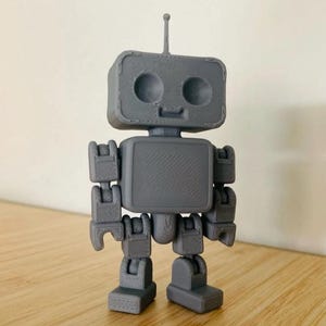 Puede incluir: Un juguete robot gris impreso en 3D con una cabeza rectangular, ojos circulares y una pequeña antena. El robot tiene brazos y piernas articulados, y un torso rectangular. El juguete está sobre una superficie de madera.