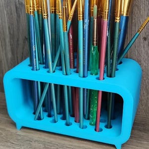 May include: A collection of paint brushes of various sizes and shapes, with gold-colored metal ferrules and colorful handles, are stored in a bright blue holder. The brushes are arranged in a vertical position, ready for use.