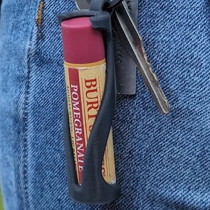 Puede incluir: Un soporte negro impreso en 3D para bálsamo labial con un bálsamo labial Burt's Bees Pomegranate. El bálsamo labial es rojo y amarillo. El soporte está sujeto a un llavero con llaves, enganchado a un bolsillo de mezclilla azul.