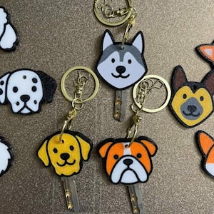 Puede incluir: Una colección de llaveros con temática de perros que presentan varias razas, incluyendo un caniche, un chihuahua, un dálmata, un husky y un bulldog. Cada llavero tiene un anillo dorado y una llave. Las caras de los perros son negras, blancas, naranjas y amarillas.