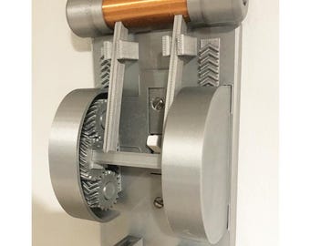 May include: A gray and copper-colored mechanical device with gears, a cylindrical copper tube, and a rectangular base. The device is mounted on a white surface and features intricate internal components.