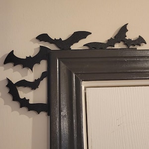 May include: Black bat decorations are affixed to the corner of a door frame. The bats are in various sizes and positions, creating a Halloween-themed accent. The door frame is dark gray, and the door is white.