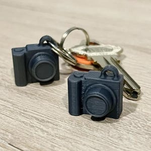 May include: Two dark blue camera-shaped keychains with a silver key ring and keys. The keychains are small, rectangular, and have a circular lens detail. The background is a light wood-grain surface.