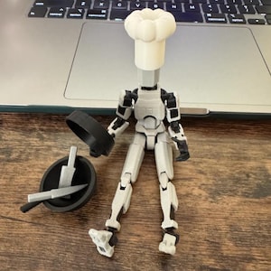 May include: A white and gray articulated figure wearing a chef's hat, holding a black frying pan and a silver knife and spatula set. The figure is posed as if cooking. The figure is on a wooden surface.