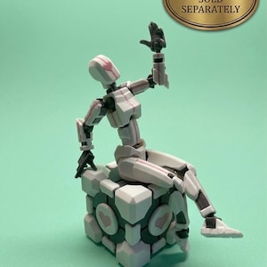 May include: A white and pink articulated action figure seated on a cube-shaped structure with heart-shaped designs. The figure is posed with one arm raised. The image includes a gold oval sign that reads "ACTION FIGURE SOLD SEPARATELY".
