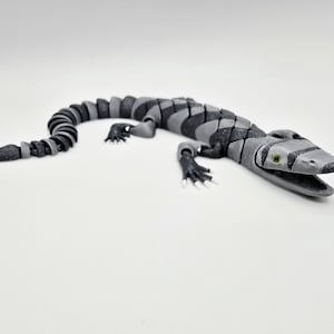 May include: A segmented, articulated alligator toy in shades of gray and black. The toy has a detailed head with open jaws and small green eyes. The body is made of interlocking segments, allowing for flexible movement. The alligator is lying on a white surface.