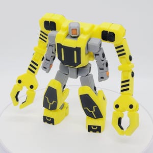 May include: A yellow and gray robot toy with articulated limbs and claw-like hands. The toy features black accents and a square head with an orange detail. The robot stands on a white surface, showcasing its mechanical design.