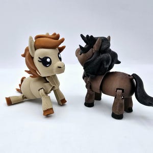 Op de afbeelding: Twee beweegbare speelgoedpaarden. Eén is beige met bruine manen en hoeven, de andere is bruin met een zwarte manen en staart. Het speelgoed is geposeerd alsof het interactie heeft. De achtergrond is effen wit.