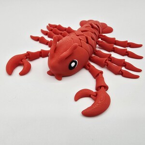 May include: A red articulated lobster toy with a segmented body, claws, and multiple legs. The toy has a detailed design and is made of a solid material. The lobster's eyes are black and white.