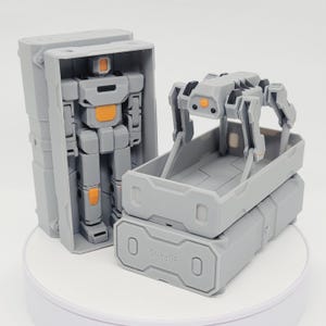 Op de afbeelding: Een grijs robotspeelgoed met oranje accenten wordt getoond naast een grijs robotachtig voertuig. De robot zit in een grijze doos. Het voertuig zit in een grijze container. De tekst "3DPRNS" staat op de container.