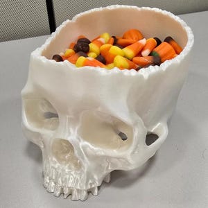 May include: A white ceramic skull shaped candy bowl filled with colorful candy corn and chocolate candies.