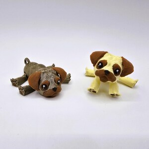 May include: Two pug dog figurines. One is lying down, the other is sitting up. Both have brown ears and patches, with black eyes and noses. The lying pug is brown and the sitting pug is yellow.