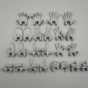 Może przedstawiać: Kolekcja małych, srebrnych modeli dłoni w różnych gestach, w tym otwarte dłonie, znaki pokoju i palce wskazujące. Dłonie ułożone są na jasnoszarej powierzchni, prezentując różnorodne pozy.