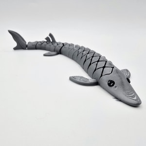 Può includere: Un giocattolo articolato grigio a forma di pesce, con pinne e un corpo segmentato. Il giocattolo è appoggiato su una superficie bianca. Il pesce ha occhi neri e un design dettagliato e strutturato.