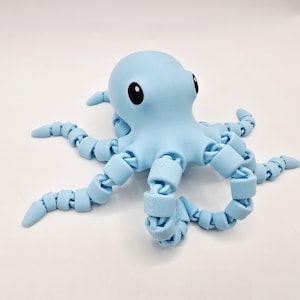 May include: A light blue articulated octopus toy with large black eyes. The octopus has a rounded head and segmented tentacles. The toy is made of plastic and sits on a white surface.