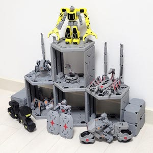 May include: A collection of grey and yellow toy robots and structures. The set includes a large yellow robot, several smaller robots, and modular grey structures with robotic creatures inside.