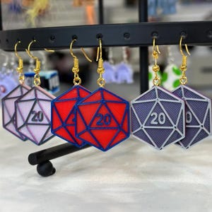 Peut inclure: Boucles d'oreilles avec un design de dé à 20 faces en rouge, violet et rose. Chaque boucle d'oreille a un crochet doré. Les dés sont numérotés avec le chiffre 20.