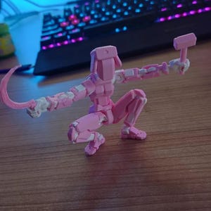 K&ouml;nnte beinhalten: Eine rosa, bewegliche Actionfigur in einer hockenden Pose, die einen Hammer und eine gebogene Klinge h&auml;lt. Die Figur hat wei&szlig;e Akzente. Der Hintergrund ist verschwommen, mit einer Tastatur sichtbar.