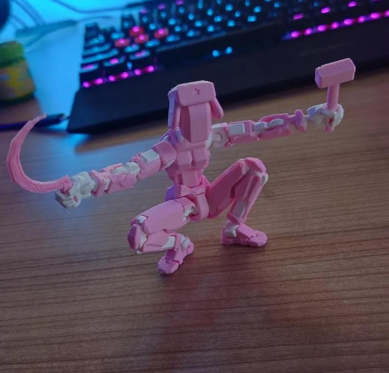 K&ouml;nnte beinhalten: Eine rosa, bewegliche Actionfigur in einer hockenden Pose, die einen Hammer und eine gebogene Klinge h&auml;lt. Die Figur hat wei&szlig;e Akzente. Der Hintergrund ist verschwommen, mit einer Tastatur sichtbar.