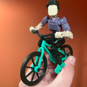 May include: A 3D printed action figure of a person riding a teal and black bicycle. The figure is wearing a black helmet and a purple shirt.