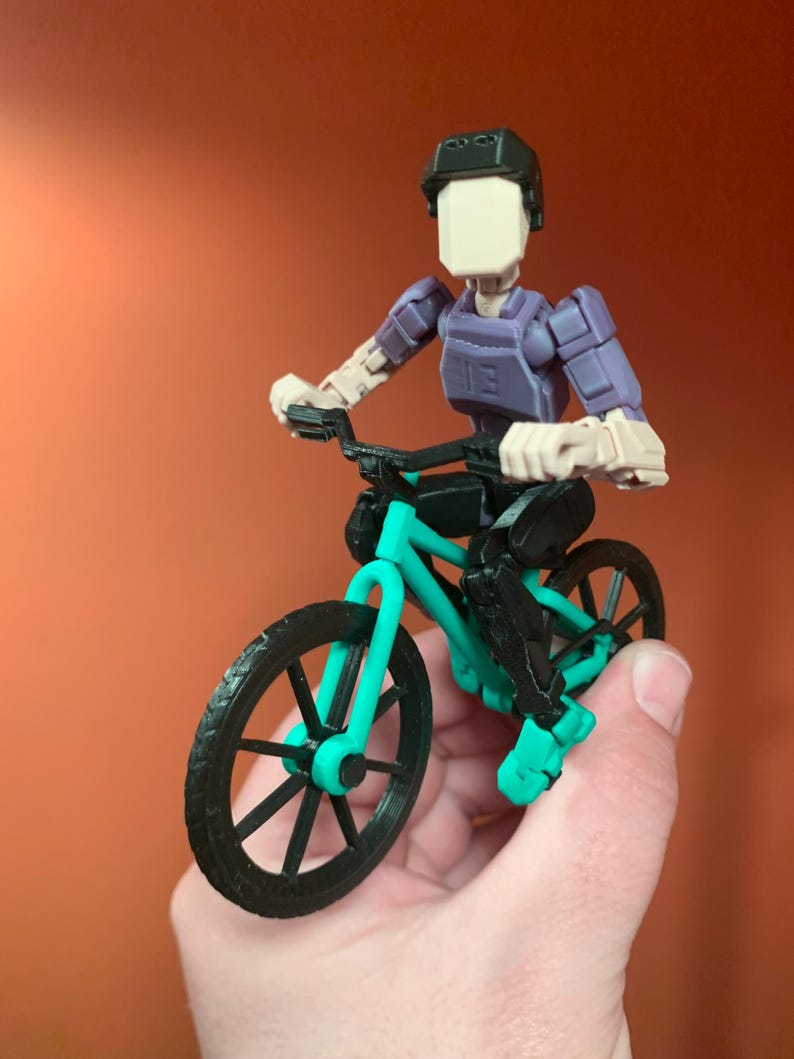 May include: A 3D printed action figure of a person riding a teal and black bicycle. The figure is wearing a black helmet and a purple shirt.