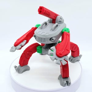 Può includere: Un robot giocattolo rosso, grigio e verde con arti articolati e un cannone. Il robot ha un corpo centrale grigio con una serie di linee verticali e due maniglie. Le gambe e le braccia sono rosse con dettagli grigi e verdi.