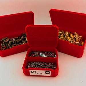Peut inclure: Trois boîtes de rangement en plastique rouge ouvertes, remplies de divers matériels métalliques. Les boîtes contiennent des inserts filetés et des rondelles de couleur laiton et argent. La boîte avant porte l'inscription "M3 A2 SS".