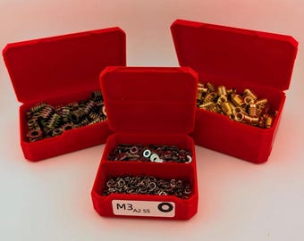 Op de afbeelding: Drie open rode plastic opbergdozen gevuld met diverse metalen hardware. De dozen bevatten messing en zilverkleurige schroefdraadinzetstukken en ringen. Op de voorste doos staat de tekst "M3 A2 SS".
