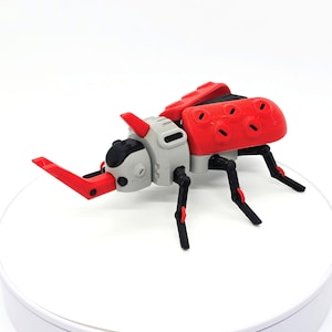 Pode incluir: Um brinquedo de besouro impresso em 3D com um esquema de cores vermelho e cinza. O besouro tem um corpo segmentado, seis pernas e grandes mandíbulas vermelhas. O design apresenta uma estética mecânica, com juntas e detalhes visíveis.