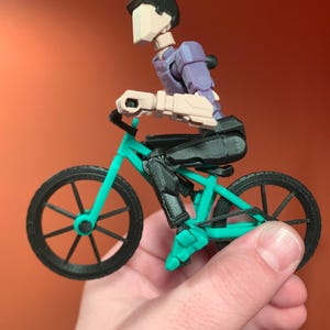 May include: A black and teal 3D printed toy bicycle with a black figure riding it. The figure has a black helmet and a purple torso.
