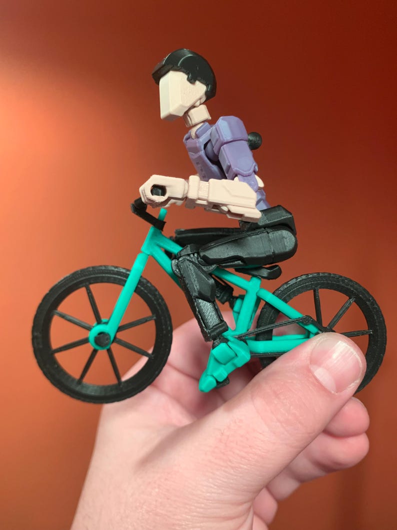 May include: A black and teal 3D printed toy bicycle with a black figure riding it. The figure has a black helmet and a purple torso.