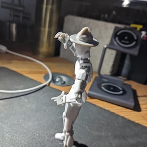 K&ouml;nnte beinhalten: Hellgraue, bewegliche Actionfigur einer Person mit Hut, die mit ausgestreckten Armen posiert. Die Figur steht auf einer dunklen Oberfl&auml;che, mit einem verschwommenen Hintergrund, der einen Computerbildschirm und andere Objekte umfasst.