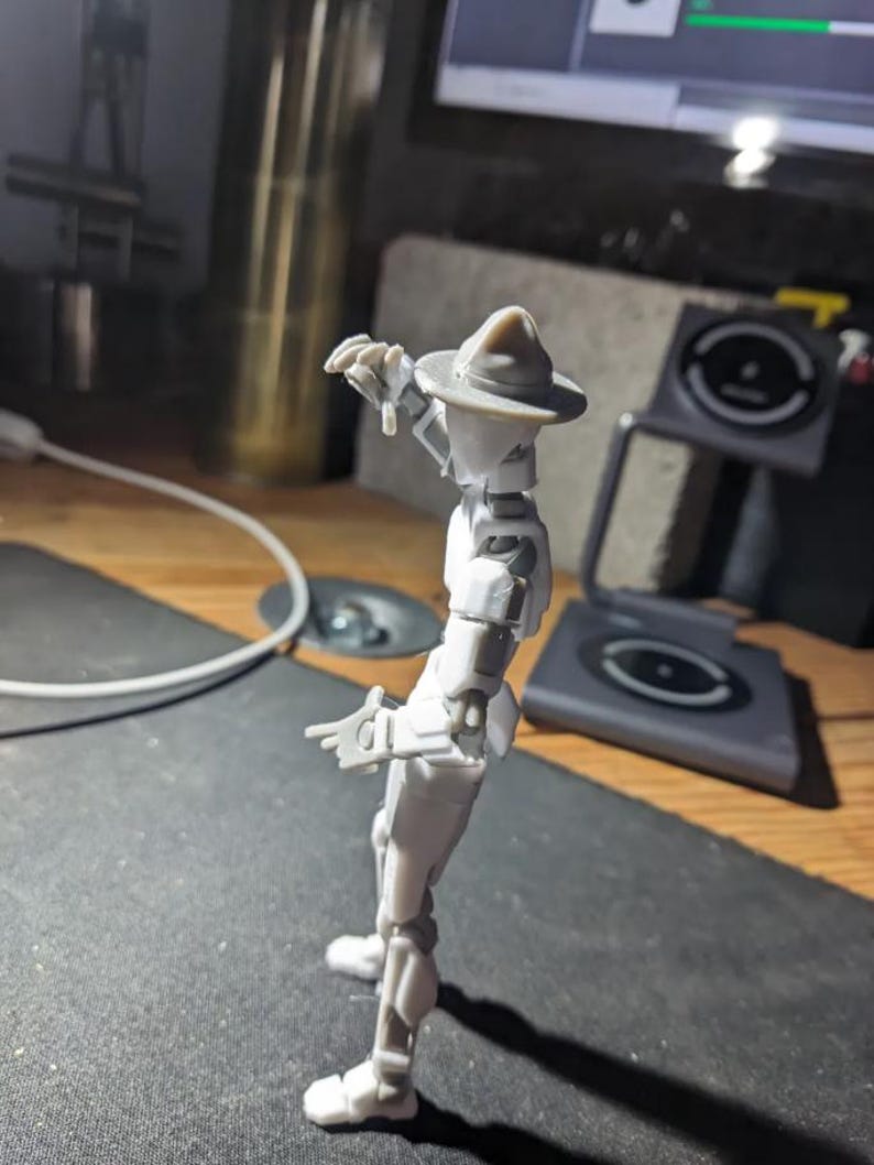 K&ouml;nnte beinhalten: Hellgraue, bewegliche Actionfigur einer Person mit Hut, die mit ausgestreckten Armen posiert. Die Figur steht auf einer dunklen Oberfl&auml;che, mit einem verschwommenen Hintergrund, der einen Computerbildschirm und andere Objekte umfasst.