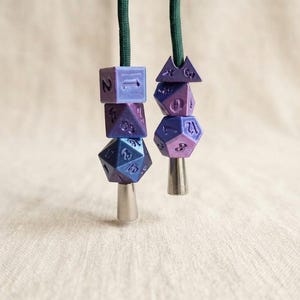 Puede incluir: Dos cordones de color verde oscuro, cada uno con una disposición vertical de tres dados de juego poliédricos de color morado y azul. Cada cordón termina en una tapa cónica de metal plateado liso. Los dados muestran varios números y formas.