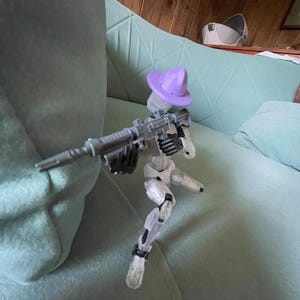 K&ouml;nnte beinhalten: Eine transparente Kunststoff-Actionfigur eines Roboters, der ein graues Gewehr h&auml;lt und einen lila Hut tr&auml;gt. Der Roboter posiert auf einem hellgr&uuml;nen Sofa.