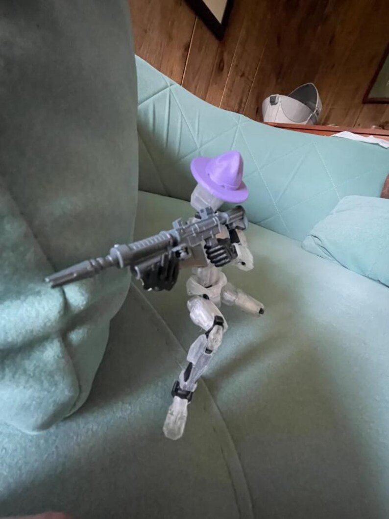 K&ouml;nnte beinhalten: Eine transparente Kunststoff-Actionfigur eines Roboters, der ein graues Gewehr h&auml;lt und einen lila Hut tr&auml;gt. Der Roboter posiert auf einem hellgr&uuml;nen Sofa.