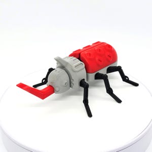 Pode incluir: Um besouro de brinquedo impresso em 3D com corpo vermelho, cabeça cinza e pernas pretas. O besouro tem um chifre vermelho grande que se estende da cabeça. O corpo apresenta recortes ovais. O brinquedo é projetado com detalhes mecânicos.