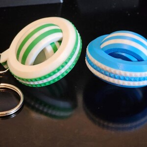 Puede incluir: Dos llaveros impresos en 3D. Uno es blanco con rayas verdes y un borde texturizado verde, sujeto a un llavero plateado. El otro es azul con rayas blancas y un borde texturizado blanco. Ambos tienen un diseño circular.
