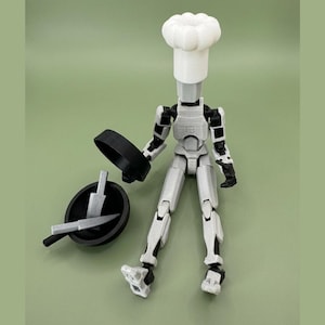 May include: A posable articulated robot figure in light gray and black, wearing a white chef hat. The figure is positioned next to a black frying pan containing two small light gray knives and a separate black lid, all rendered in a matte finish.