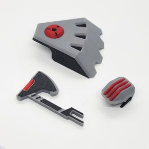 Op de afbeelding: Een verzameling 3D-geprinte grijze en zwarte componenten met rode accenten. De set bevat een schildachtig stuk, een kleine bijl en een afgerond object met drie rode strepen. De onderdelen zijn gerangschikt op een witte ondergrond.
